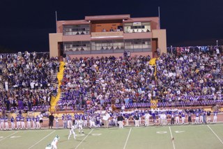 The stadium at night during a football game with the bleachers filled with sports fans.