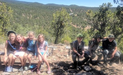 Summer camp kids sitting and being silly on benches with mountains scenery. Hiking with the summer camp kids on one of the camp days, up at Abbey Alley benches, with SMNHC property and buildings down below.