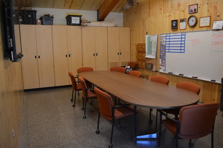Conference room with 9 orange chairs around brown oblong table, tall wood cabinets, white board, and t.v. screen