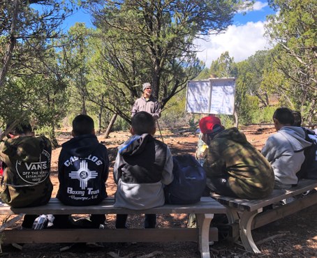 Instructor standing by white board outside with students facing him sitting on benches