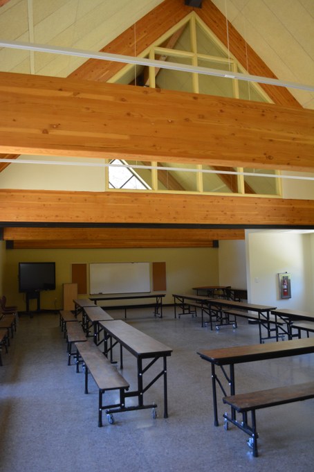 Large multi-purpose room with cafeteria-style benches and tables, and triangle wooden beam feature halfway up wall to ceiling