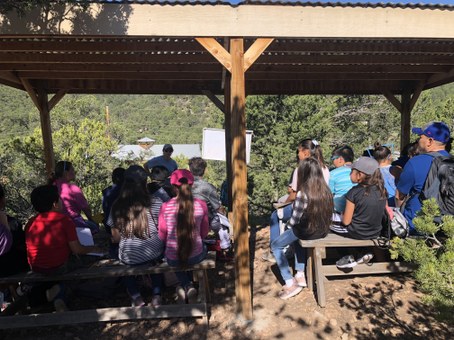 Students sitting on benches under shade structure facing instructor, with building and trees across the way