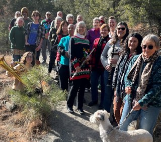 A large group of people, some holding instruments, and one dog, standing outdoors with trees around.